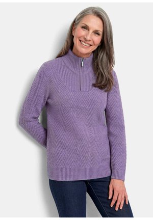 Lächelnde Frau, die einen violetten Reißverschluss-Pullover mit Chevron-Strickmuster und dunkelblaue Jeans vor neutralem Hintergrund trägt.