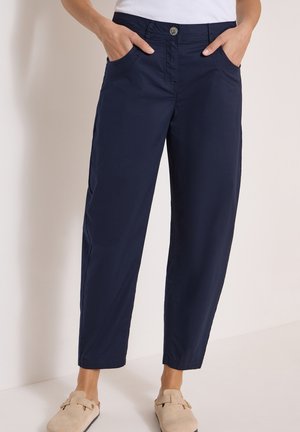 Woman wearing navy blue straight-leg pants with front pockets and beige slip-on shoes standing against a light background.
