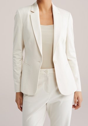 Femme portant un blazer blanc sur mesure par-dessus un top côtelé beige et un pantalon blanc taille haute assorti avec une fermeture par bouton.