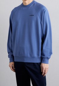 Blå sweatshirt med rund hals och raglanärmar. Enkel design med en liten svart logotyp nära halslinningen. Mjuk, slät textur.