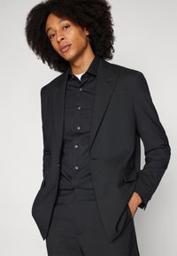 Black suit with a tailored jacket and matching trousers. The shirt is also black with buttons. Smooth texture, slim fit design.