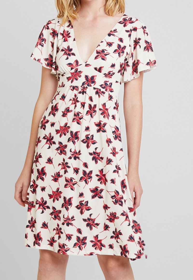 Femme portant une robe crème avec de courtes manches flottantes et un imprimé floral rouge et noir, avec un profond décolleté en V et une taille cintrée.