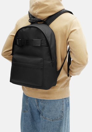 Person trägt einen beigefarbenen Hoodie und blaue Jeans mit einem schwarzen Rucksack, der eine Fronttasche und verstellbare Träger hat.