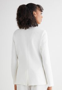 Witte op maat gemaakte blazer met een enkele split aan de achterkant, lange mouwen en een gladde textuur, die het minimalistische ontwerp en de schone lijnen benadrukt.