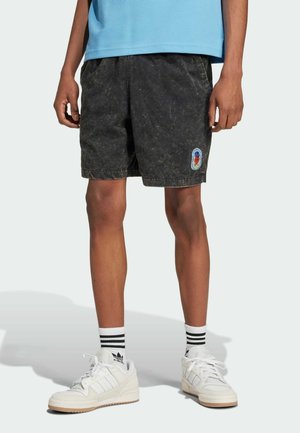 Osoba nosząca czarne, wypłukane szorty z małą naszywką, białe buty sportowe Adidas i białe skarpetki z czarnymi paskami, stojąca na jednolitym tle.