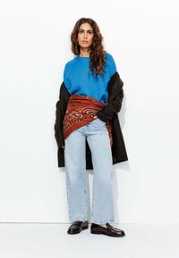 Sweatshirt bleu, manteau oversized marron, étole à motifs paisley rouges, jean large bleu clair et chaussures marron. Textures lisses avec une coupe décontractée.
