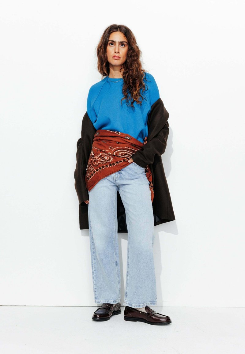 Sweatshirt bleu, manteau oversized marron, étole à motifs paisley rouges, jean large bleu clair et chaussures marron. Textures lisses avec une coupe décontractée.