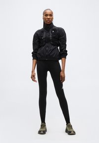 Ψηλή γυναίκα με μαύρο αθλητικό μπουφάν, μαύρα κολάν με λογότυπο και ελιάς πράσινα αθλητικά παπούτσια, στέκεται μπροστά σε απλό φωτεινό φόντο.