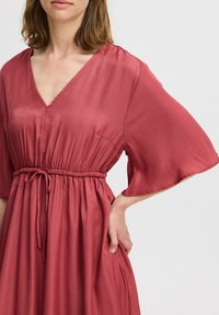 Robe rose en tissu lisse et fluide. Elle présente un décolleté en V, des manches courtes en forme de cloche, et une taille élastique avec un lien décoratif.