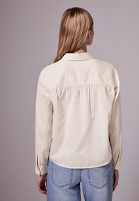 Crème-kleurig shirt met lange mouwen, een kraag en een geplooid achterdetail, gecombineerd met lichtblauwe jeans. Zachte, gladde stoftextuur.
