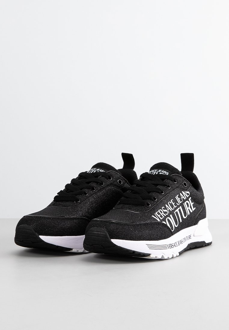 Un par de zapatillas Versace Jeans Couture en blanco y negro con cordones negros y texto de marca en los laterales y en la suela, exhibidas sobre una superficie gris.