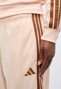 Gros plan d’une main reposant sur le côté, portant un pantalon rose clair et une veste avec des triples rayures marron et des détails de logo.