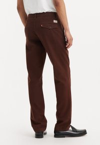 Pantaloni din twill brown cu croială dreaptă, având o buzunare din spate cu nasture și un mic etichetă a mărcii. Textură netedă, purtați cu mocasini negri.