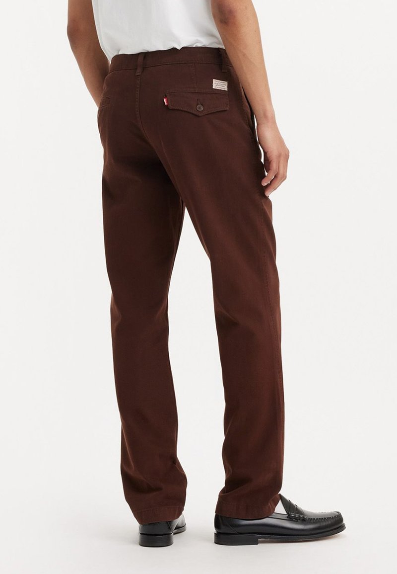 Pantaloni in twill marrone con taglio dritto, dotati di una tasca posteriore con bottone e una piccola etichetta del marchio. Texture liscia, indossati con mocassini neri.