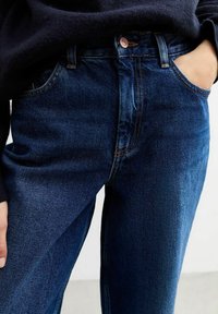 Jeans in denim blu scuro con vita alta, design a gamba dritta e un leggero sbiadimento. Presentano cuciture arancioni e tasche frontali classiche.