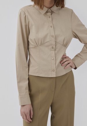 Frau trägt beige langärmliges Hemd mit Knöpfen und Kragen sowie olivgrüne Hose, Hand in der Hüfte, vor schlichtem grauem Hintergrund.