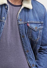 Giacca in denim con colletto in shearling, cuciture a vista e chiusure con bottoni in argento. Indossata sopra una maglietta grigia a girocollo.