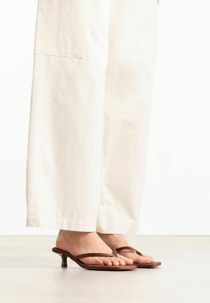 Voeten met bruine teenslippers met lage hak, gecombineerd met wijde off-white broekspijpen tegen een effen achtergrond.