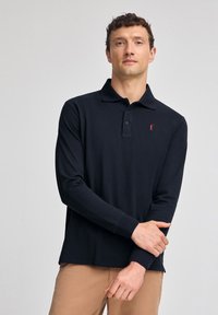 Polo a maniche lunghe di colore blu navy con colletto, presenta una chiusura con tre bottoni e un piccolo emblemma rosso sul petto. Tessuto dalla texture morbida.