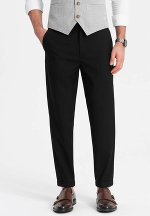 Pantalones negros a medida con una textura suave, corte ajustado y puños enrollados, combinados con zapatos de cuero marrón.