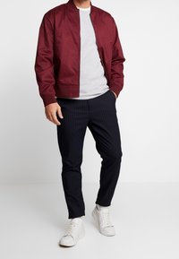 Burgundy bomberjacka över en grå T-shirt, kombinerad med marinblå randiga byxor och vita sneakers; mjuk textur och avslappnad passform.