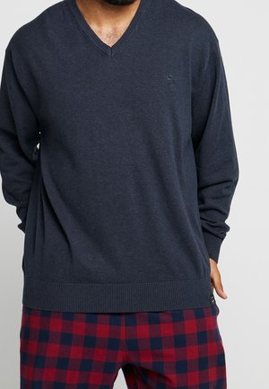 Hombre que lleva un jersey de cuello en V azul marino oscuro y pantalones a cuadros rojos y negros, de pie con una mano detrás de la espalda.