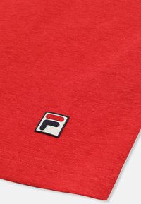 Gros plan sur un tissu rouge avec un petit logo rectangulaire près du bord, présentant un "F" stylisé en rouge et noir.