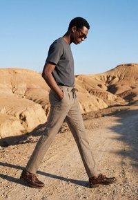 Homme portant des lunettes de soleil, un t-shirt gris, un pantalon à carreaux et des chaussures marron marchant sur un chemin de terre dans un paysage sec et rocailleux sous un ciel clair.