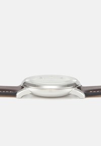 Boîtier de montre en acier inoxydable argenté, forme ronde, avec un verre lisse. Bracelet en cuir marron foncé avec des accents de couture contrastants.