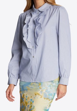 Mujer que lleva una blusa azul clara con rayas finas y volantes en la parte delantera, y una falda colorida con estampado abstracto.
