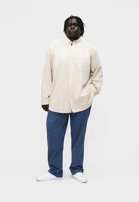 Volwassene met een donkere huid en dreadlocks staand, gekleed in een beige oversized overhemd met strepen, blauwe broek en witte sneakers tegen een effen achtergrond.