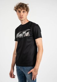 Camiseta negra con un estampado gráfico que presenta las palabras "LES HOMMES" en blanco y plateado. Está hecha de una tela suave, tiene mangas cortas y un cuello redondo.