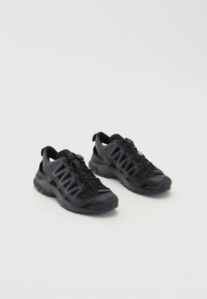 Black athletic shoes with a mesh upper, rubber sole, and elastic laces. Features a reinforced toe and distinct side panel design.