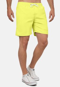 Pantaloni da bagno giallo neon con vita elastica, coulisse bianca e un accento di marca. Tessuto liscio e design lungo fino al ginocchio.