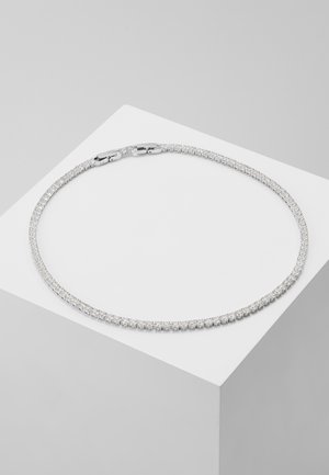 Collier tennis rond en argent avec de petites pierres scintillantes présenté sur un piédestal géométrique blanc sur fond gris.