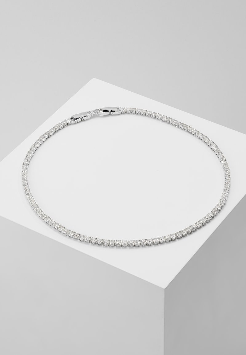 Collier tennis rond en argent avec de petites pierres scintillantes présenté sur un piédestal géométrique blanc sur fond gris.