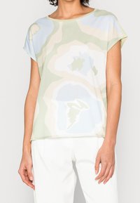 Blouse à manches courtes en tissu léger avec un motif floral pastel pâle. Elle présente un col rond et une coupe décontractée, dans des tons verts, bleus et crème.
