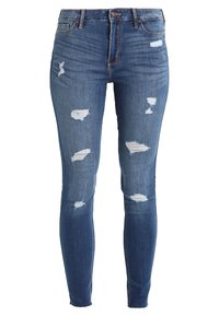 Vaqueros ajustados en denim azul medio, con múltiples parches desgastados, un diseño entallado y un estilo clásico de cinco bolsillos con trabillas para el cinturón.