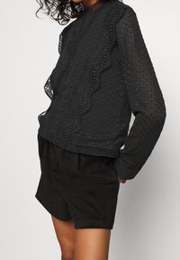 Musta tekstuuriga ülasärk, millel on täppide muster, pitsaktsendid, pikad varrukad ja kõrge kaelus, koos mustade lühikeste pükstega, millel on lõdva lõige.