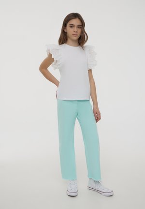 T-shirt bianca con maniche in pizzo arricciate, abbinata a pantaloni verde menta e sneaker bianche. Sfondo semplice, modella in piedi con una mano sui fianchi.