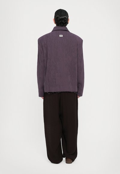 Übergroße lila Jacke mit strukturiertem Finish, ausgestattet mit einem Kragen und einem unvollendeten Saum, kombiniert mit locker sitzenden dunkelbraunen Hosen.
