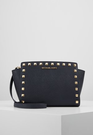 Czarny nadgarstek Michael Kors z złotymi kwadratowymi ćwiekami obrysowującymi przód i odłączanym paskiem na nadgarstek, na jednolitym tle.