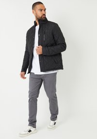 Veste matelassée noire avec un col montant, fermeture éclair et fermeture à pressions. Portée par-dessus un t-shirt blanc, associée à un pantalon gris et des baskets blanches.