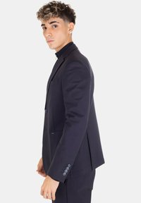 Giovane uomo con capelli ricci che indossa un completo blu scuro, in piedi di profilo su uno sfondo bianco semplice.