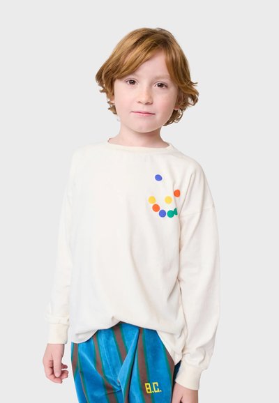 Bijela majica dugih rukava s šarenim točkastim uzorkom na prsima; nosi se s plavim i smeškim prugastim hlačama; mekani materijal, opušteni kroj.