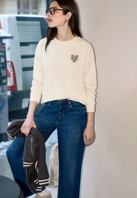 Femme portant un sweat crème avec un cœur imprimé léopard, jean bleu, lunettes, tenant une veste foncée, debout à l'intérieur près d'une fenêtre avec un arrière-plan urbain.