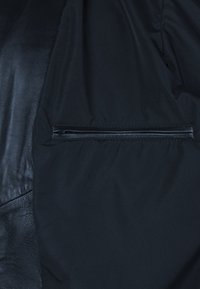 Schwarze Jacke mit einer Kombination aus mattem Stoff und glänzenden Lederelementen, ausgestattet mit einer Reißverschlusstasche auf der Vorderseite. Insgesamt glatte Textur.