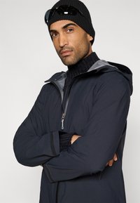 Sort isoleret jakke med grå accenter, der har lynlås foran og hætte. Båret over en chunky sort rullekrave. Tilbehør inkluderer en sort beanie og solbriller.