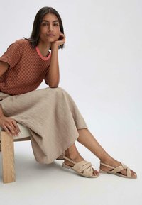 Beige knoten Sandalen mit flacher Sohle, kombiniert mit einem hellbeigen Leinenrock und einem braun gestreiften Hemd, sitzend auf einem Holzstuhl.