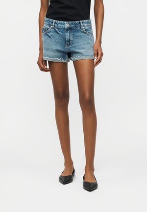 Kék farmer rövidnadrágot viselő, karcsú lábú személy, a szegély szakadt, fekete hegyes orrú lapos cipőben és fekete felsőben, egyszínű háttér előtt.